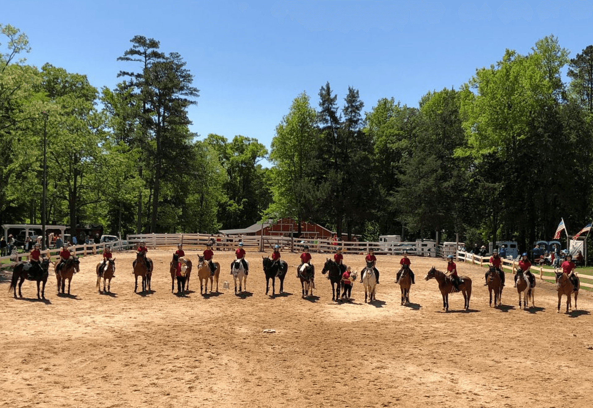 Group of riders on horseback lined up in a sandy equestrian arena surrounded by trees.