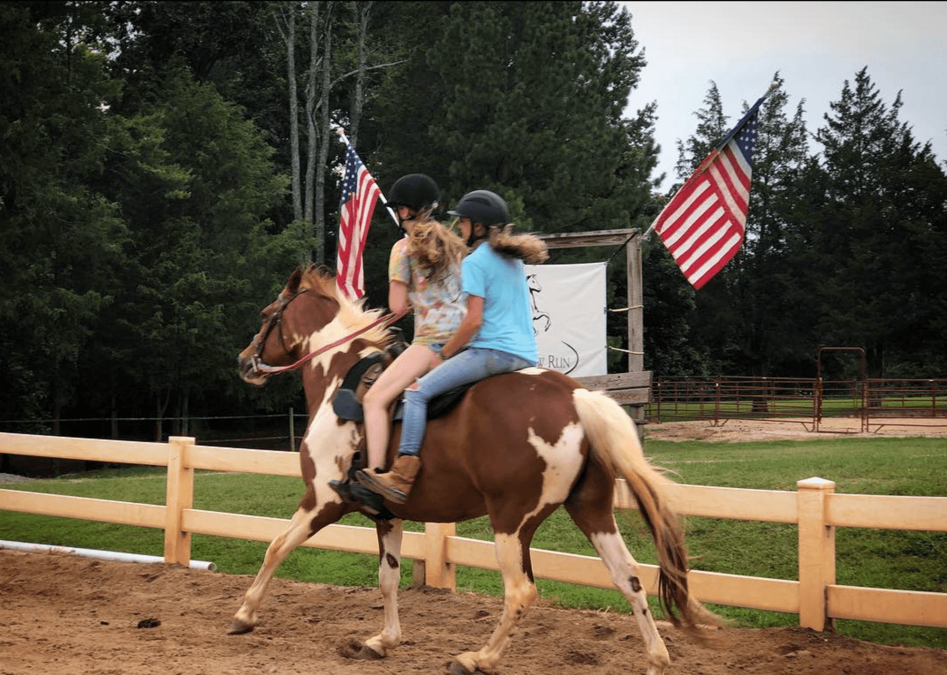 Two people riding a horse in an arena with American flags and trees in the background.